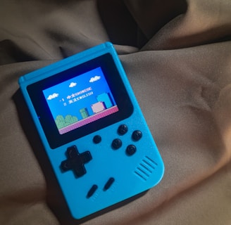 A handheld gaming device with a bright blue casing rests on a soft, dark fabric background. The screen displays a pixelated video game scene with options to select either Chinese or English language. The layout includes a classic D-pad and buttons, reminiscent of retro gaming consoles.